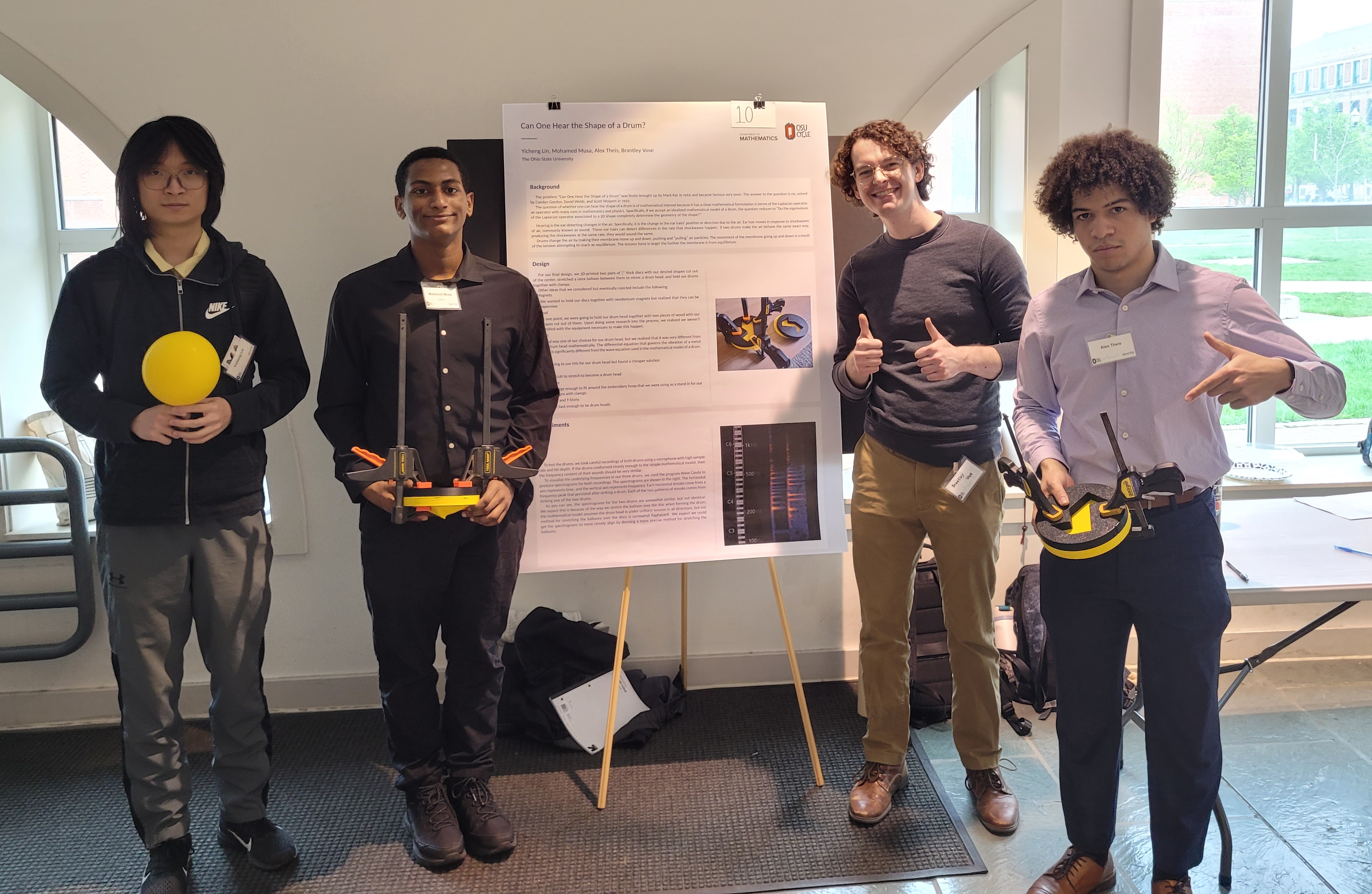 Four people holding the drum prototypes, standing in front of a conference poster.